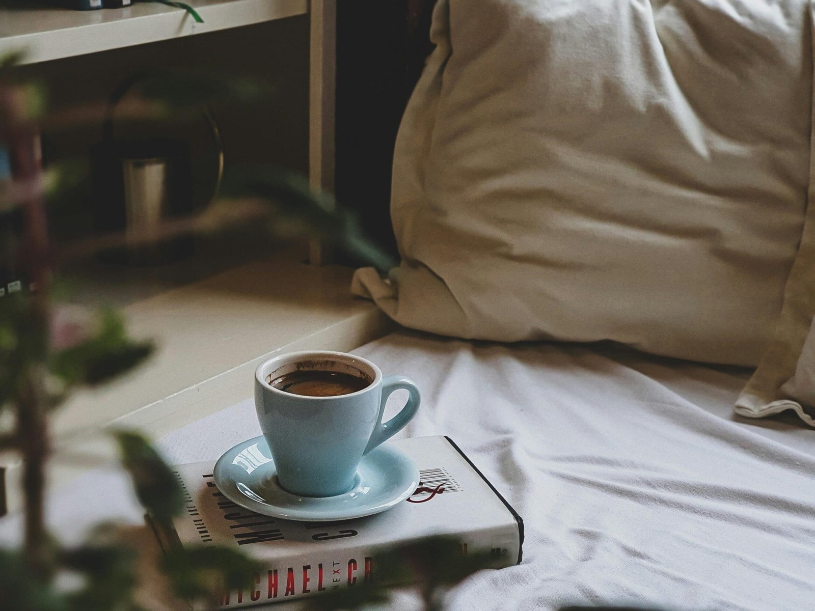 A comforting scene featuring a warm cup of coffee resting on a book, set on a cozy bed by a bookshelf.